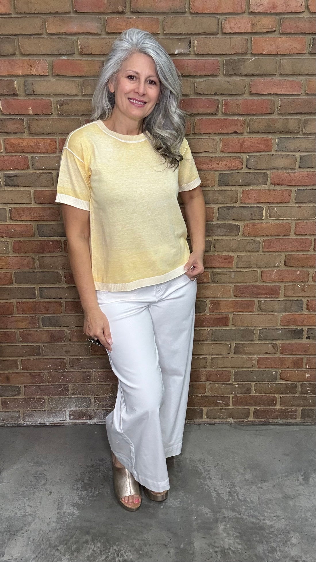 Woman wearing the Sunshine Short Sleeve Sweater, a yellow sweater and the White Twill Cropped Flare Leg pants standing against a brick wall.