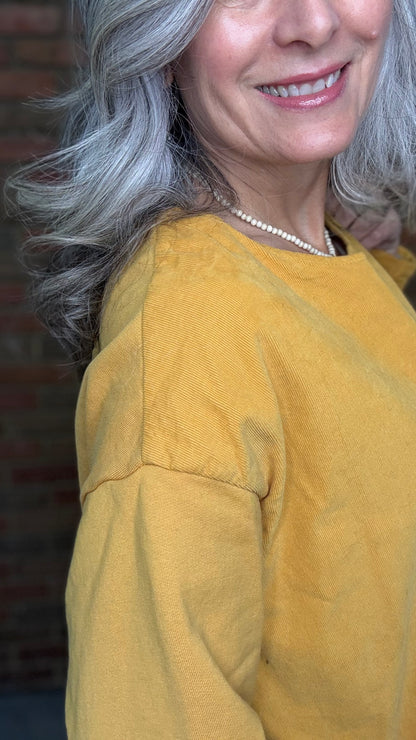 Woman wearing the Solid Mustard Puckered Pullover  and the Upside Down Jeans standing in front of a brick wall.