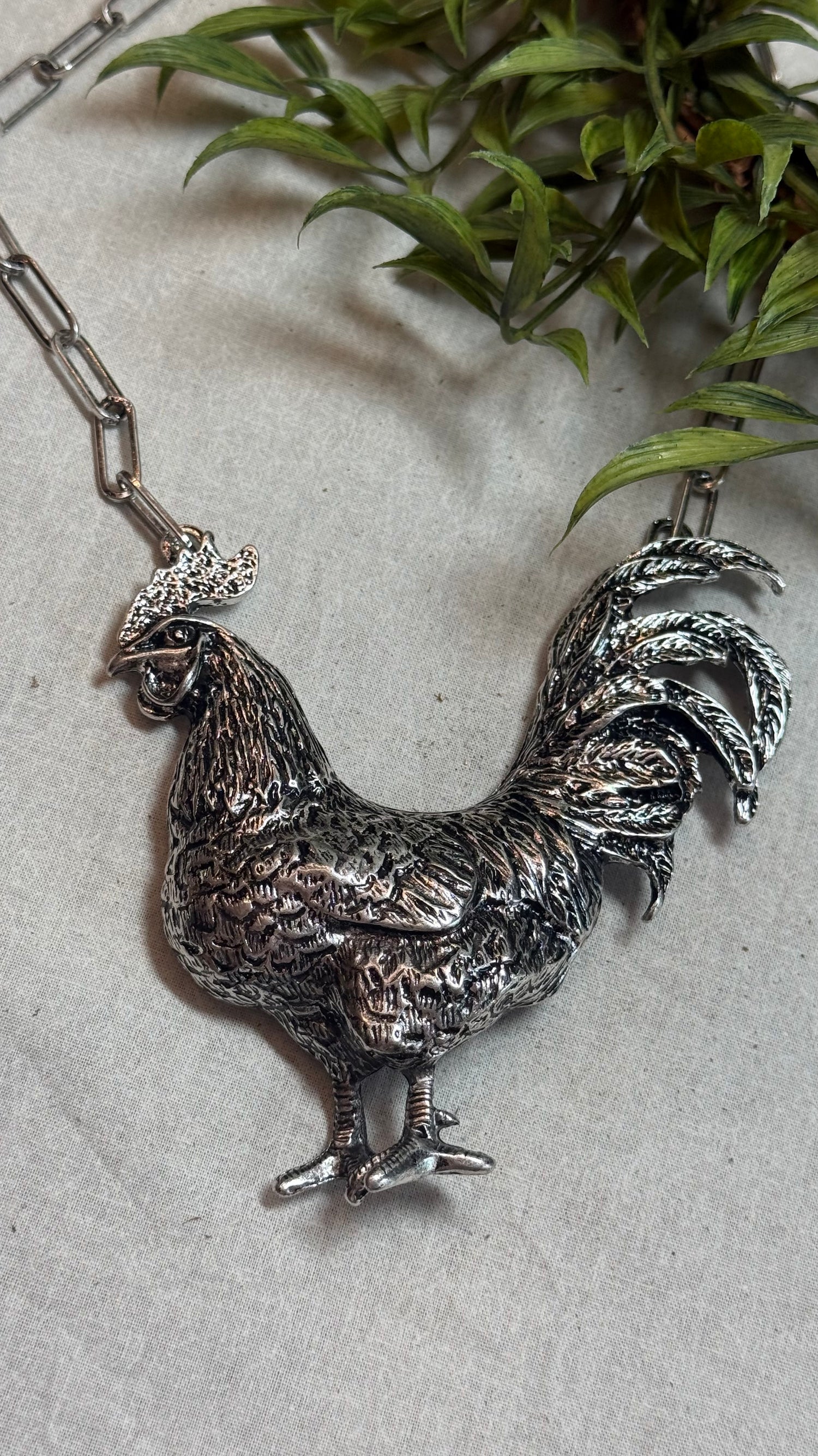 A detailed silver rooster pendant necklace on a silver link chain, displayed on a light gray textured surface with green leaves in the background.