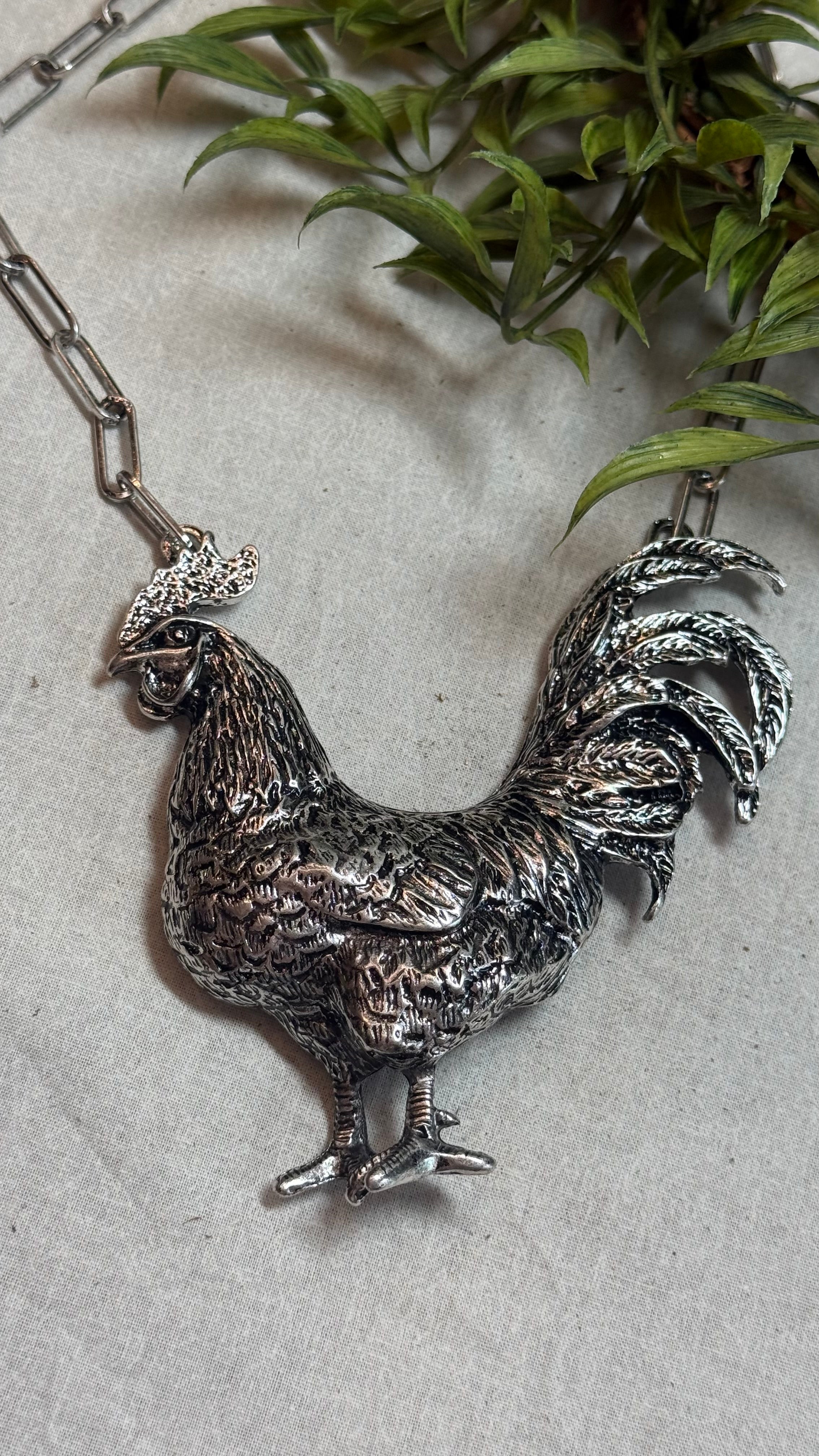 A detailed silver rooster pendant necklace on a silver link chain, displayed on a light gray textured surface with green leaves in the background.