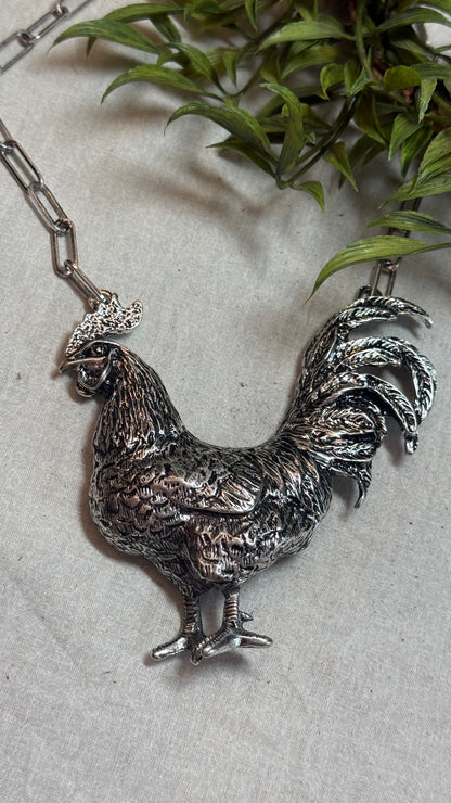 A detailed silver rooster pendant necklace on a silver link chain, displayed on a light gray textured surface with green leaves in the background.