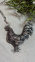 A detailed silver rooster pendant necklace on a silver link chain, displayed on a light gray textured surface with green leaves in the background.