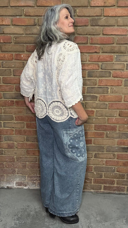 Woman wearing a white lace top and blue denim pant with floral patterns against a brick wall.