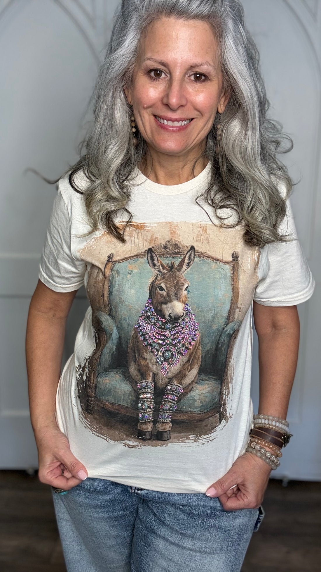 A smiling woman with long gray hair models a white graphic t-shirt featuring a detailed illustration of a donkey wearing a crown and colorful patterned accessories, seated on a blue chair. She is also wearing blue jeans and bracelets.