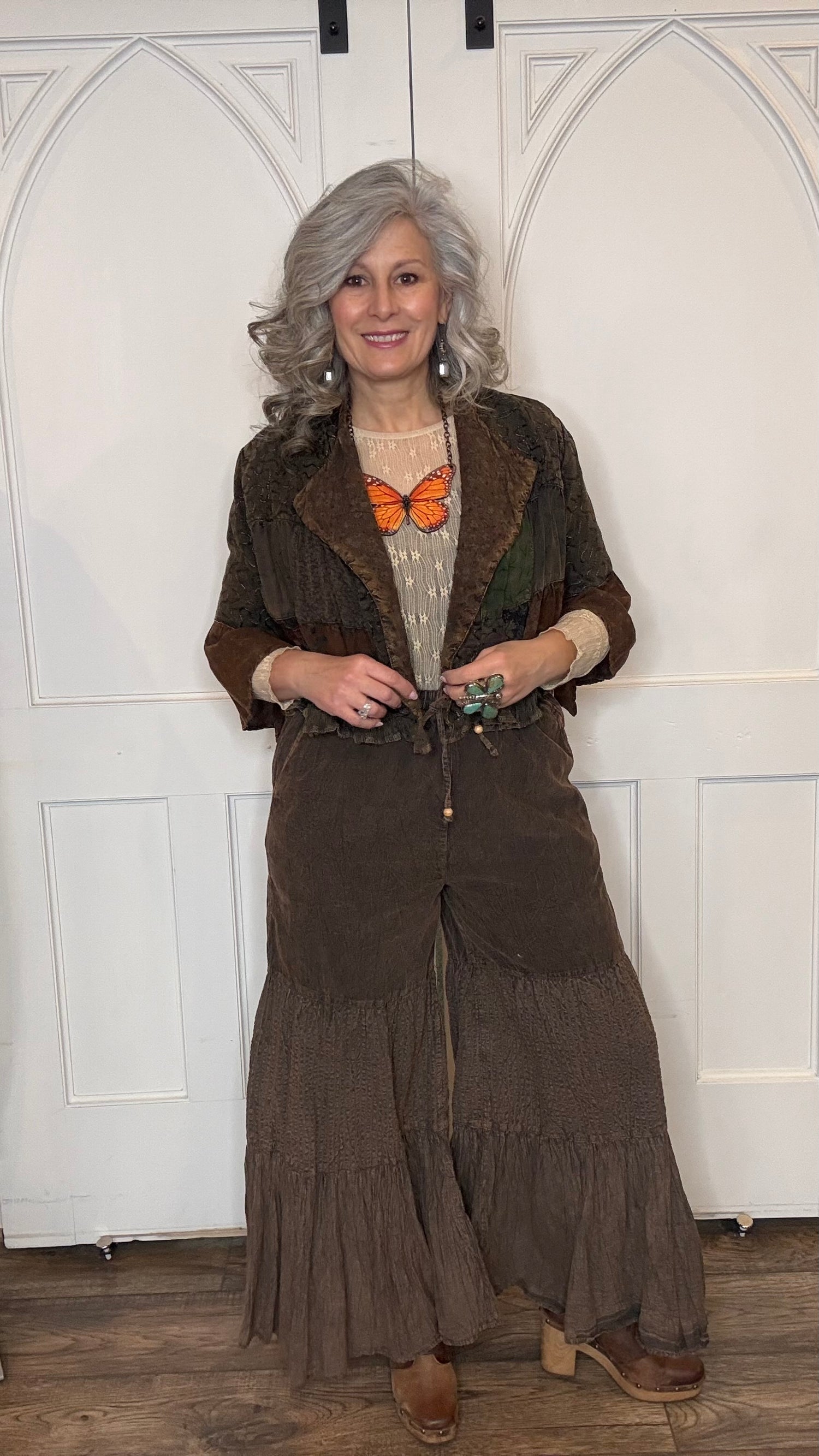 Woman wearing Boho Embroidered Lapel Crop Jacket, beige lace top with Monarch Butterfly Necklace and Amber Boho Chic pants against a white paneled wall.