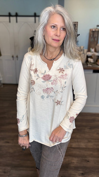A woman with silver hair wearing a cream-colored long-sleeve top with pink and blue floral embroidery, paired with gray pants, standing in a home interior setting.