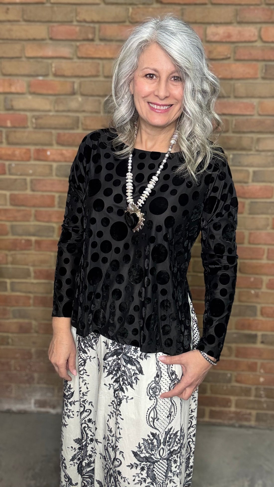 Woman wearing Black Velvet Burnout Top, Floral Print Wide Leg Pant against a brick wall. 