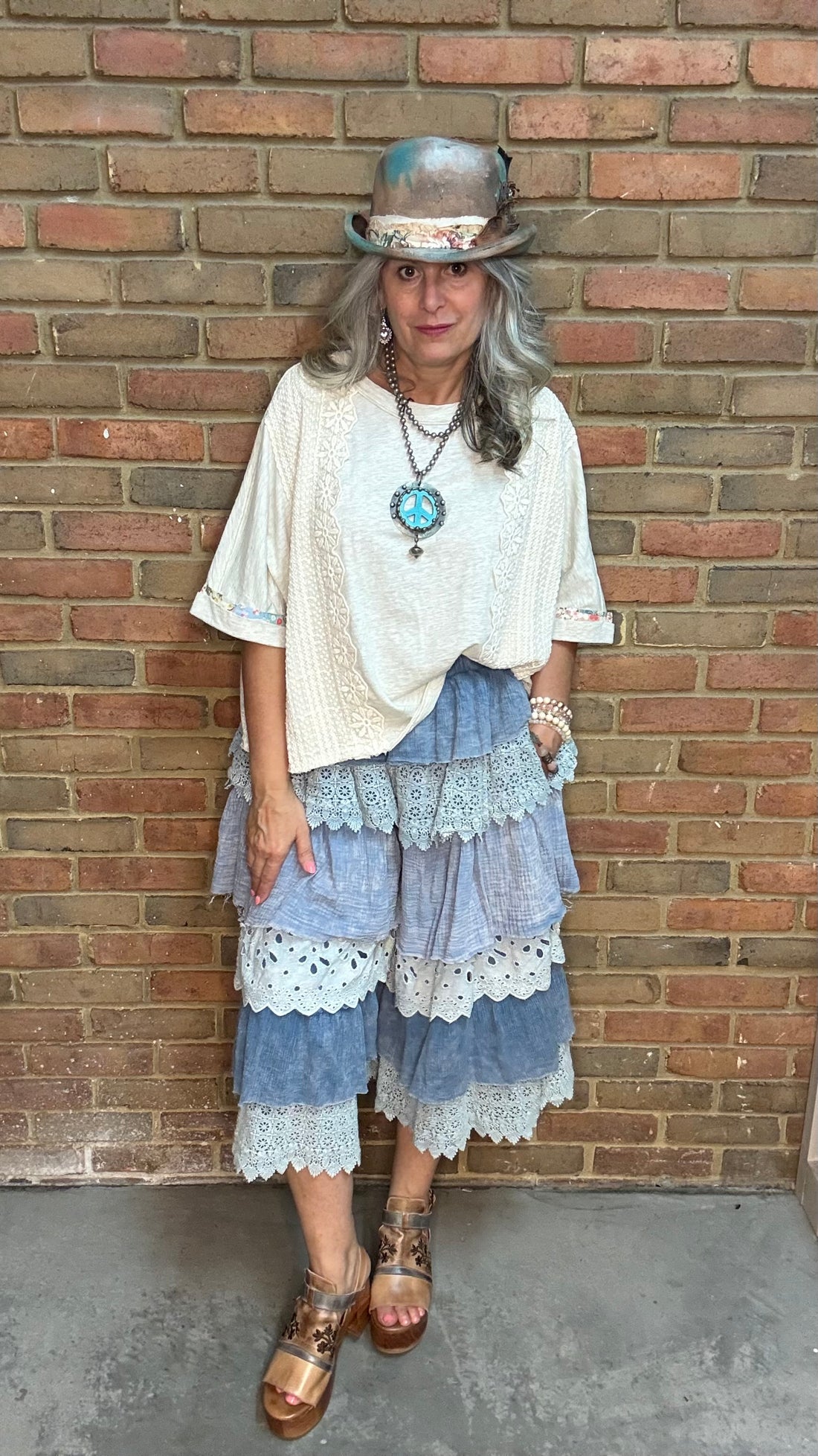 A woman wearing a bohemian-style outfit consisting of a cream short-sleeve top, a blue and white tiered pant with lace trim, a hand painted hat, and brown sandals, standing against a brick wall.
