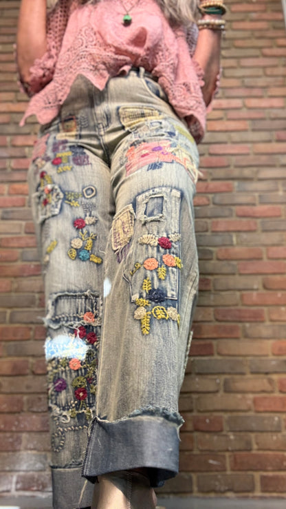 A person wearing heavily embellished light-wash denim jeans with colorful floral embroidery, patchwork applique, and distressed details, paired with a pink lace top, standing against a red brick wall.