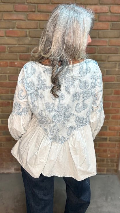 A woman with long gray hair is shown from the back wearing a light blue-colored blouse with blue-gray floral embroidery and dark blue jeans, standing in front of a brick wall.