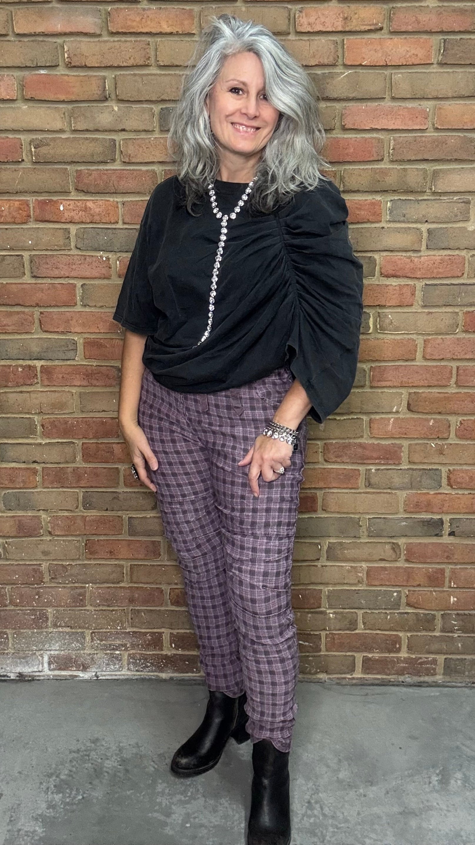 Person wearing a black top and purple checkered pants standing against a brick wall.