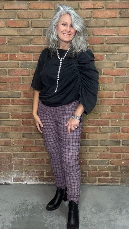 Person wearing a black top and purple checkered pants standing against a brick wall.