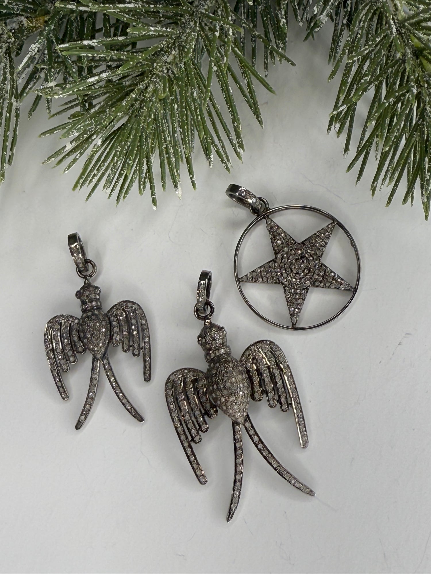 Pave diamond pendants with star and bird designs on a white surface with greenery.