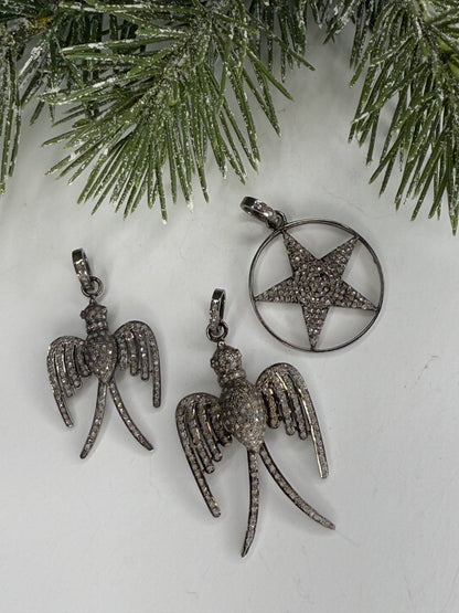 Pave diamond pendants with star and bird designs on a white surface with greenery.