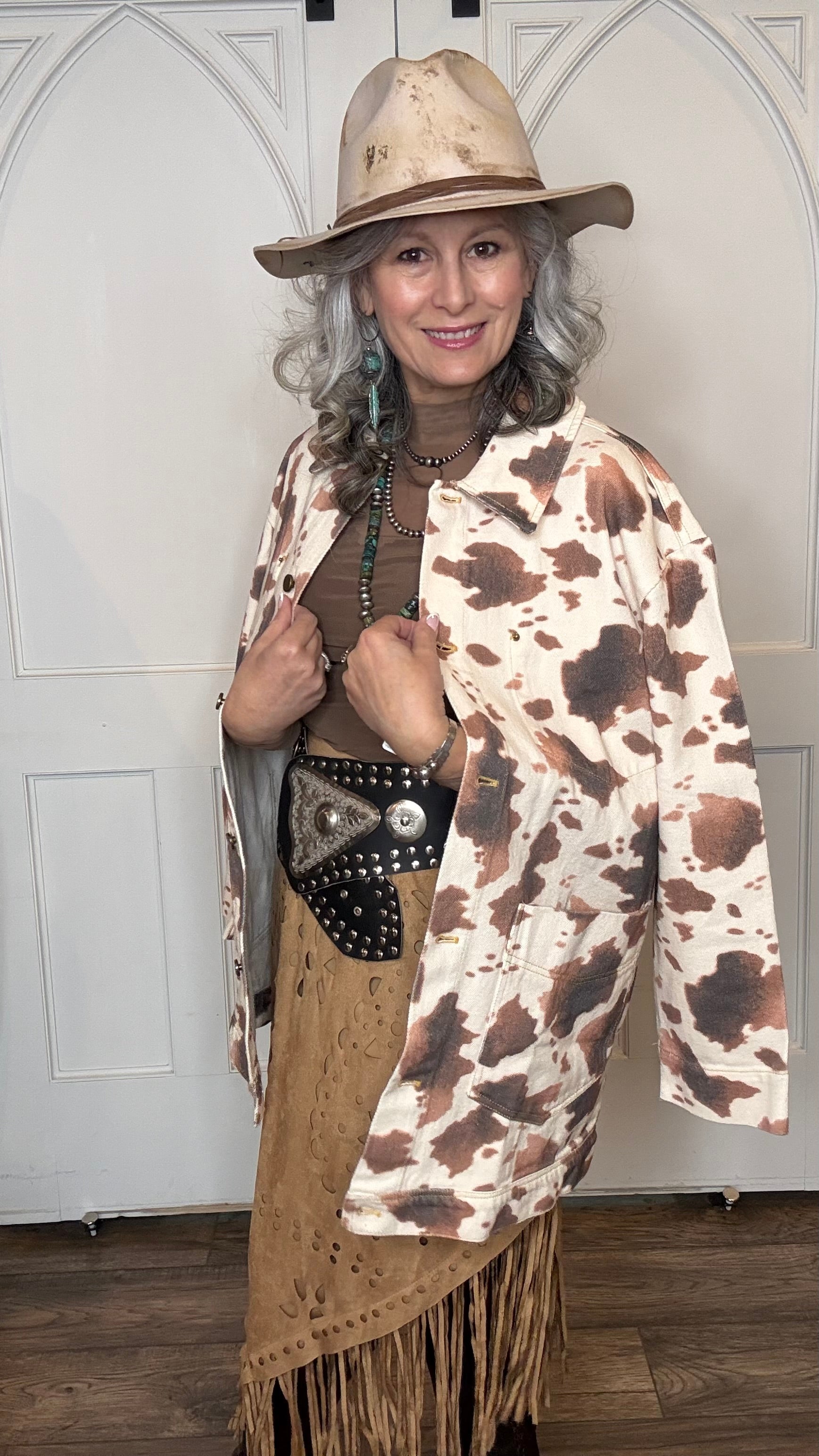 Woman wearing the Cow Print Denim Jacket, the Camel Faux Suede Fringe Skirt, topped the solid Mesh top in Brown and the Wide Triangle Buckle Belt.  Shoes are the Lane Plain Jane Boots, and she is wearing the Pop&