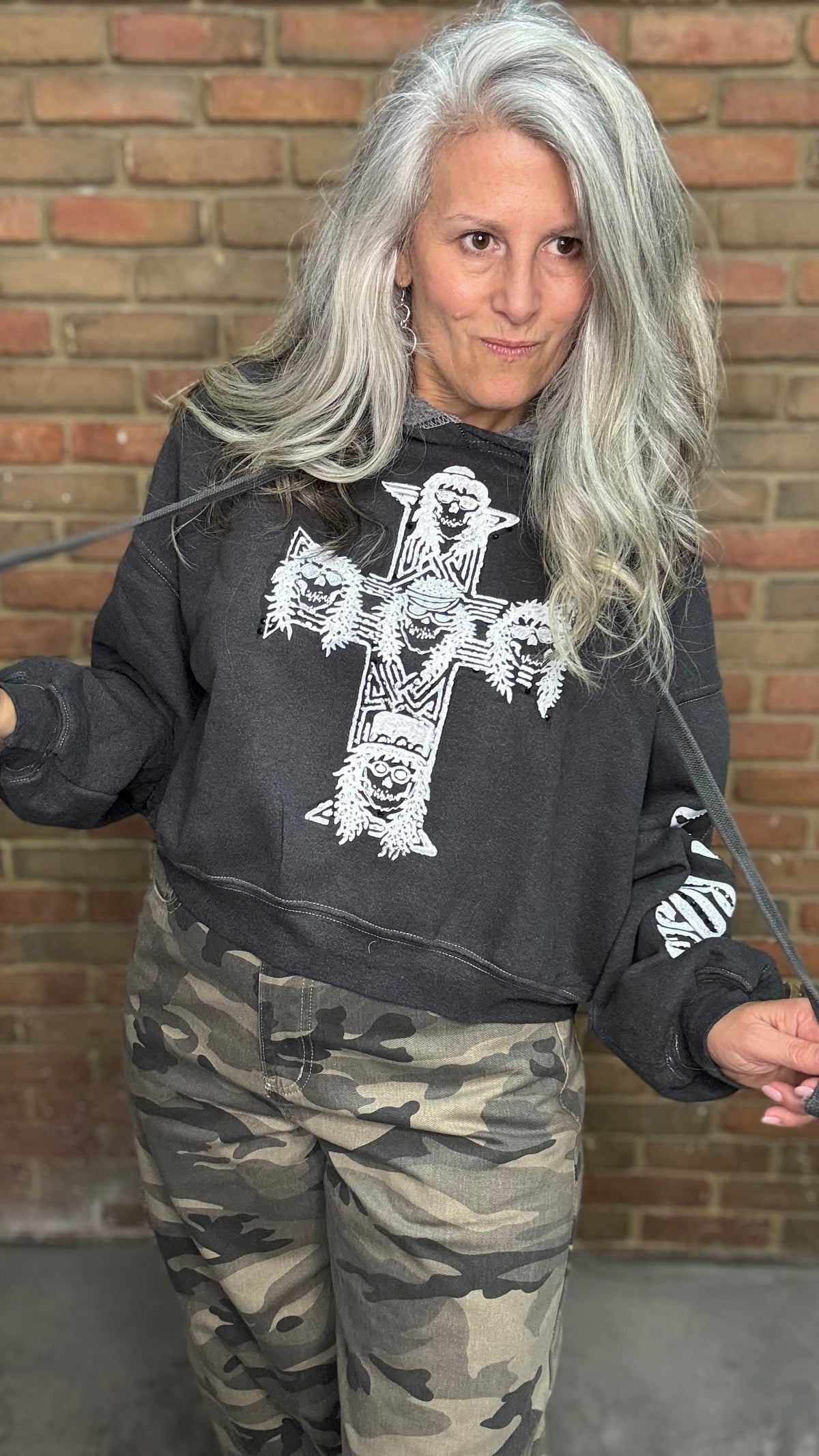 A woman with long silver hair wearing a black graphic sweatshirt with a white cross design and camouflage pants, standing in front of a brick wall.