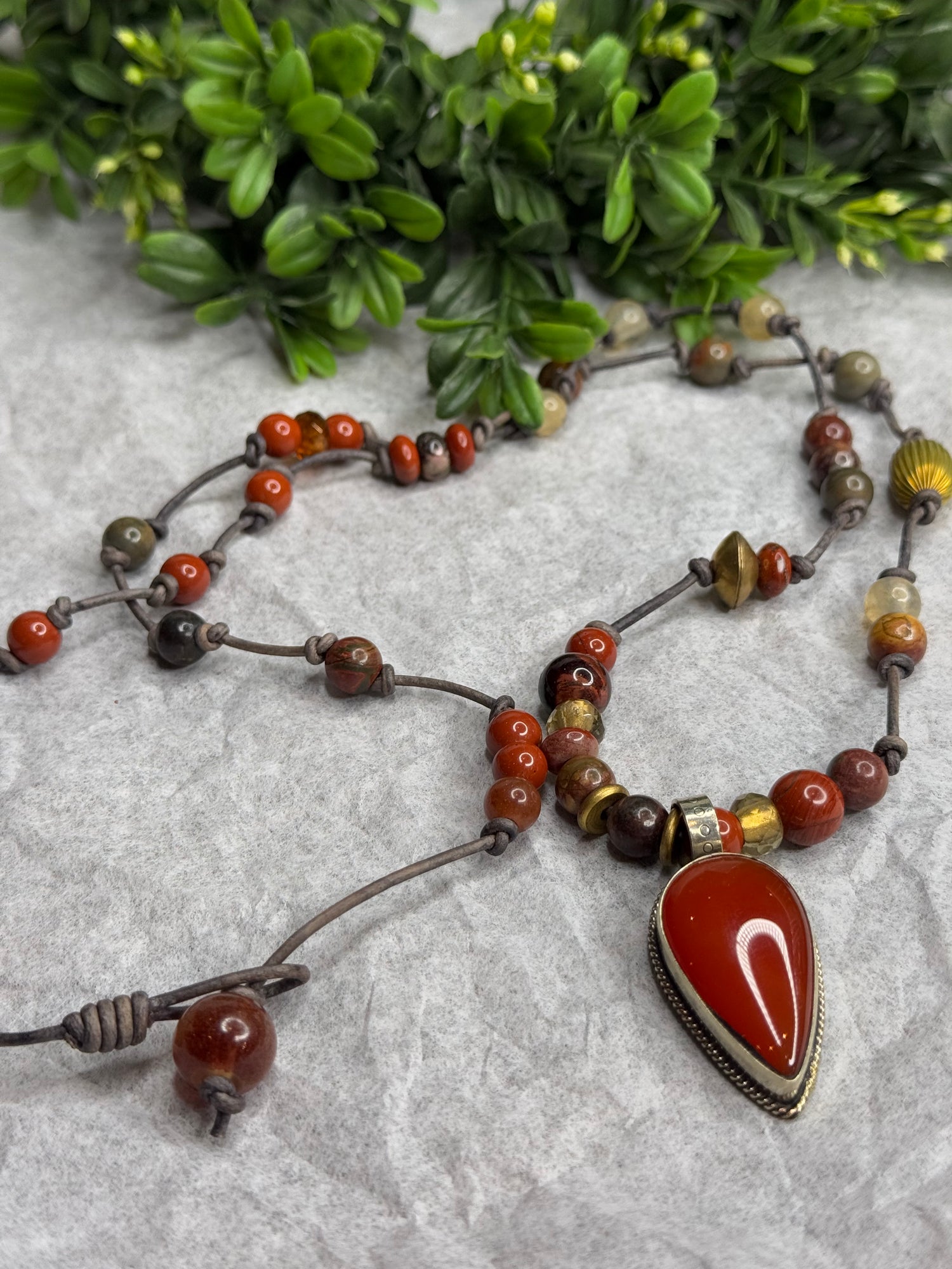 Beaded necklace with a large red stone pendant on a textured surface with greenery.