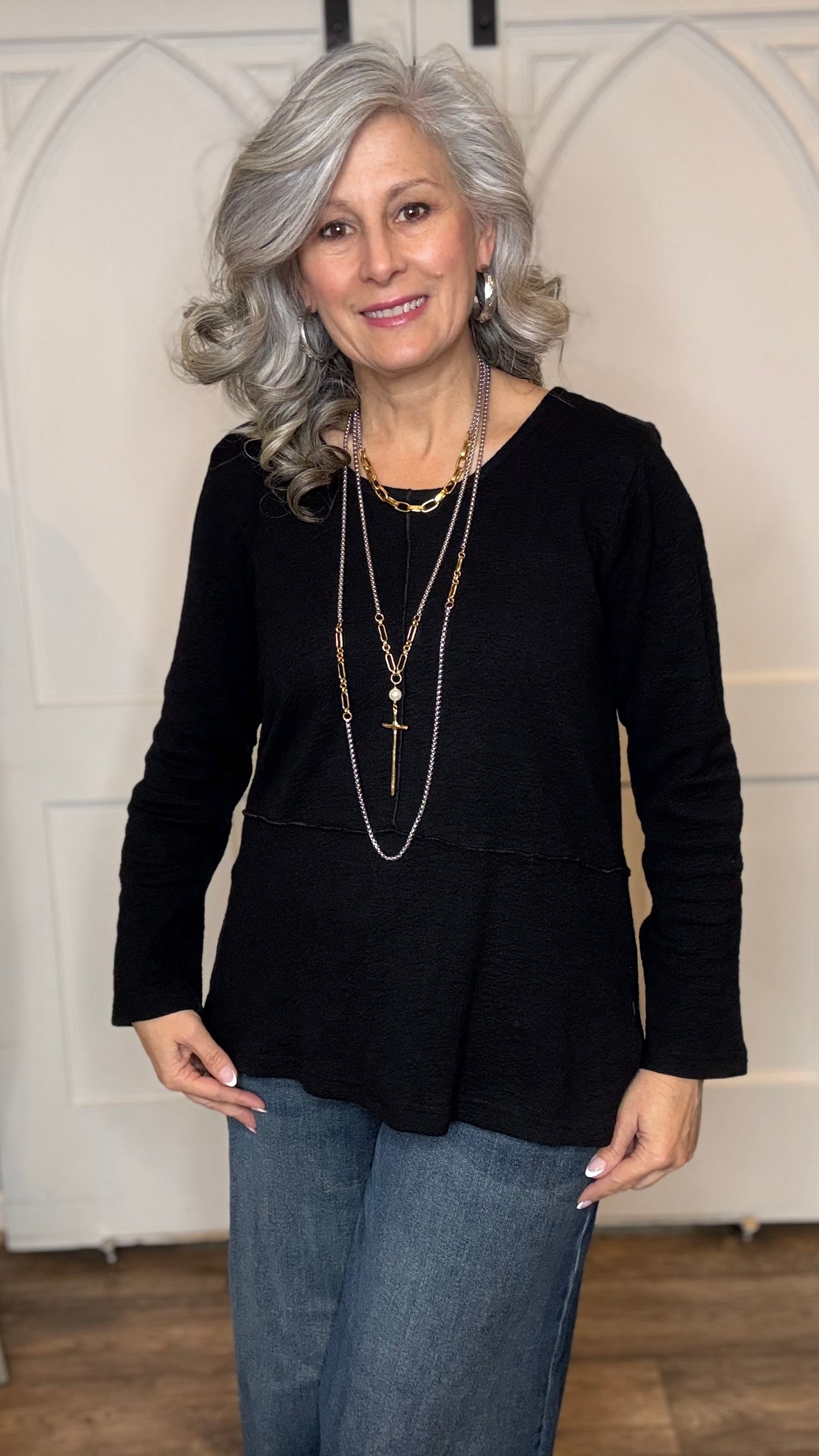 Woman wearing The Black Wanda Round Neck Top layered with the Silver Snake and Gold Paperclip Chain Necklace, the Silver Snake and Gold Paperclip Chain Choker Necklace and the Gold Cross w/Pearl Necklace  paired with  Judy Blue Palazzo jeans indoors against a white paneled wall.