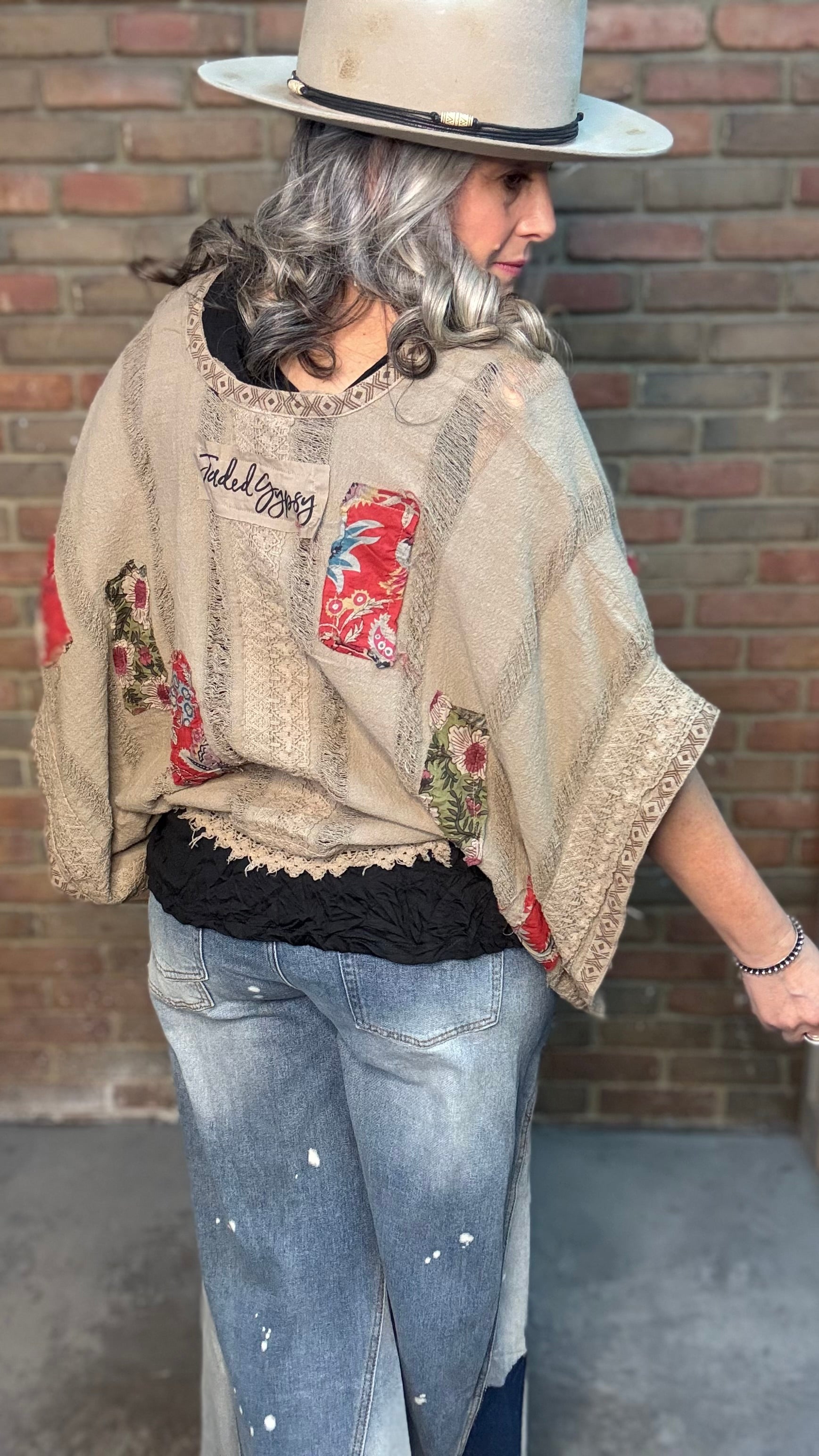 Woman wearing Garden Drift Top with a hat, jeans, and belt against a brick wall.