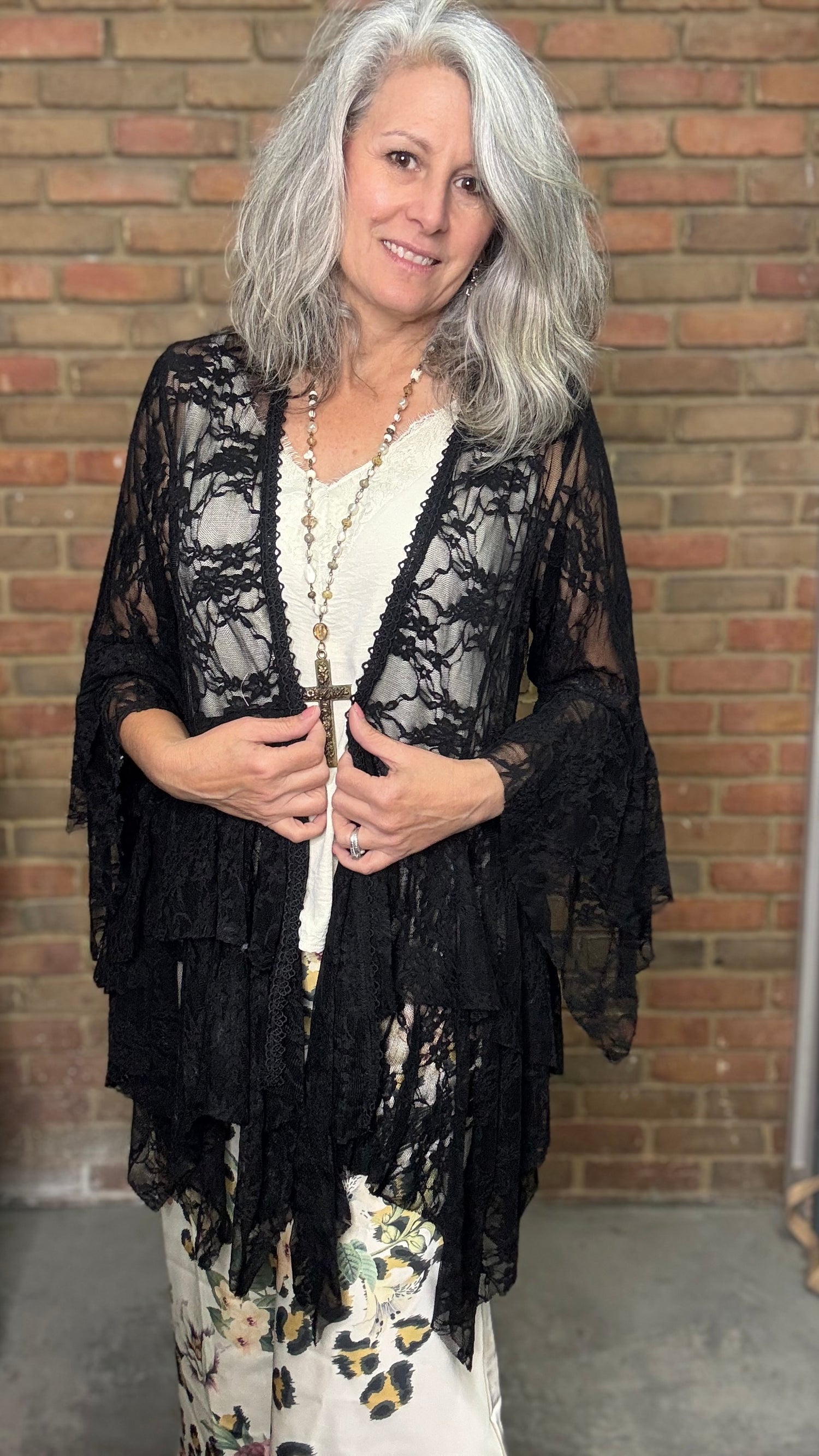 Woman wearing a black lace cardigan over a floral dress against a brick wall.