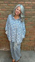 A woman with gray hair stands against a brick wall wearing a loose light blue floral button-up shirt and light gray jeans with white embroidery.