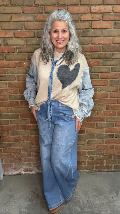 Woman wearing a sweater with a heart design and jeans standing against a brick wall.