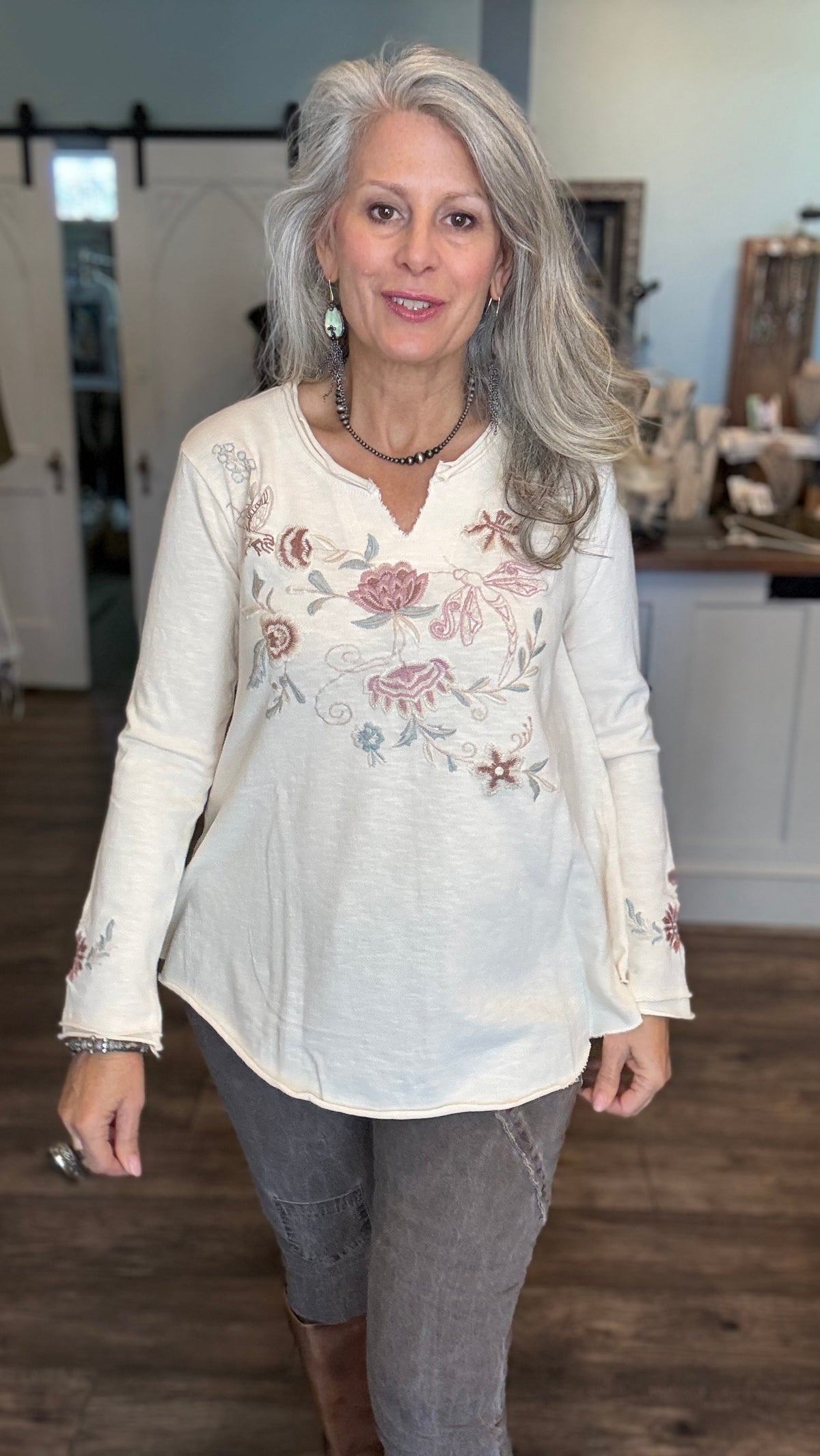 A woman with long gray hair wearing a cream-colored long-sleeve top with pink and blue floral embroidery, paired with gray pants, standing in a rustic indoor setting.