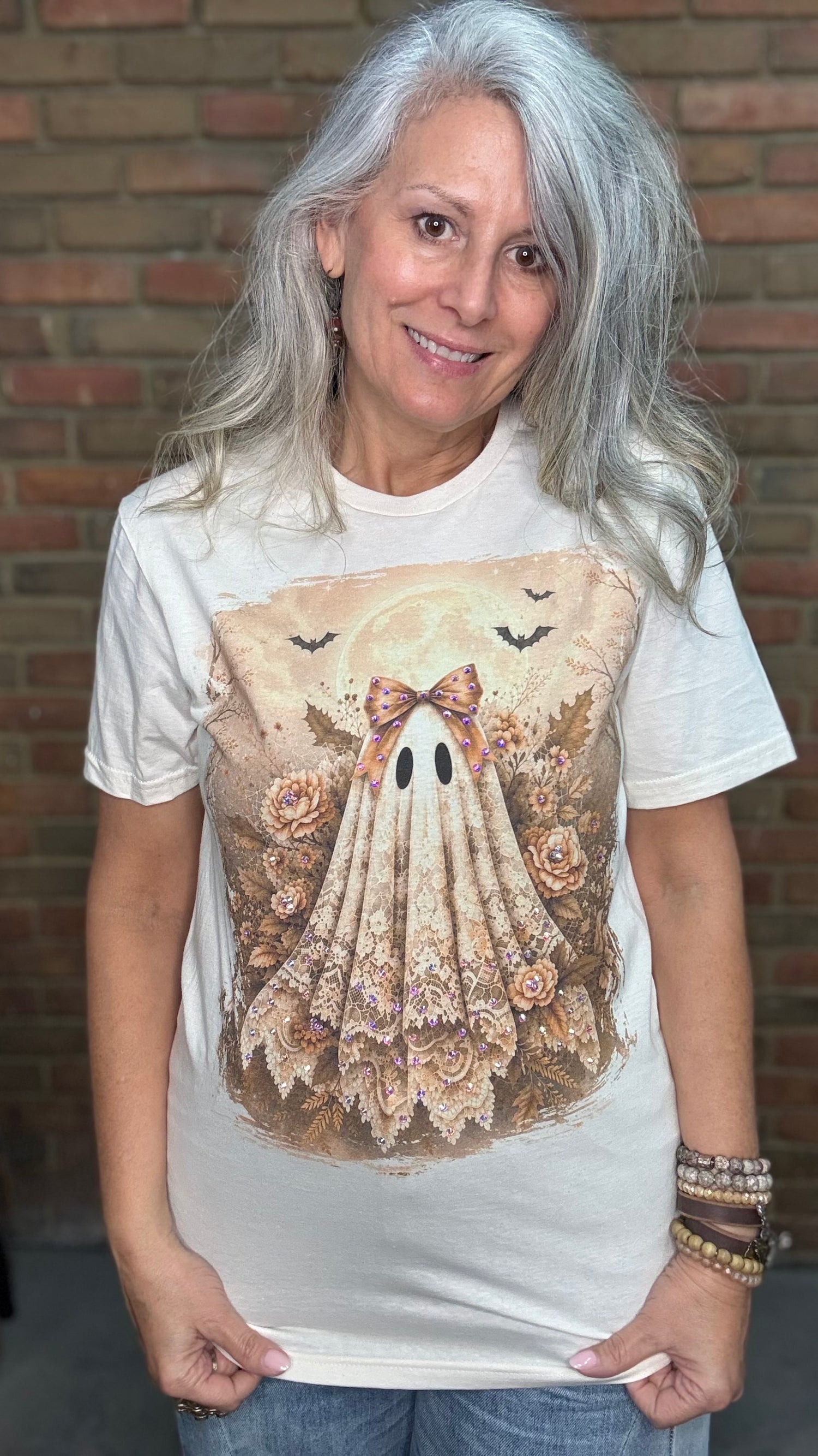 A woman with long gray hair wearing a cream-colored graphic t-shirt with a detailed illustration of a ghostly figure made of lace and roses, posed in front of a brick wall.