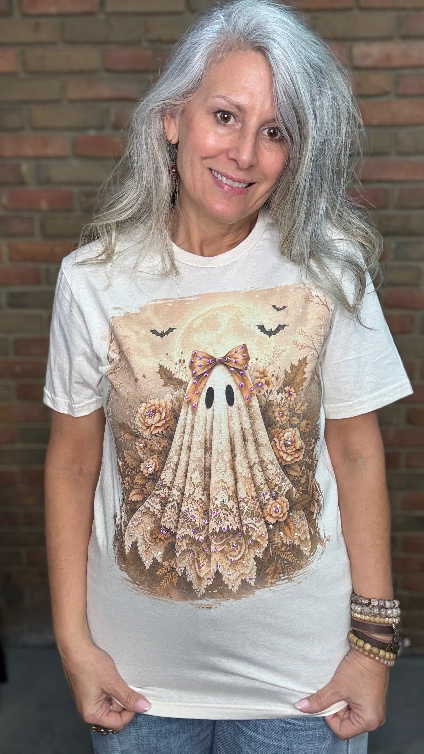 A woman with long gray hair wearing a cream-colored graphic t-shirt with a detailed illustration of a ghostly figure made of lace and roses, posed in front of a brick wall.