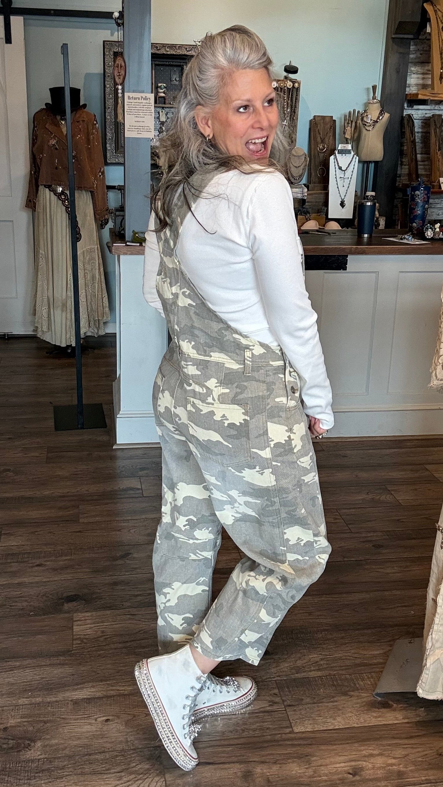 A woman with blonde hair tied back is wearing a pair of camouflage print overall jumpsuits over a white long-sleeve shirt and white sneakers, standing inside a boutique.