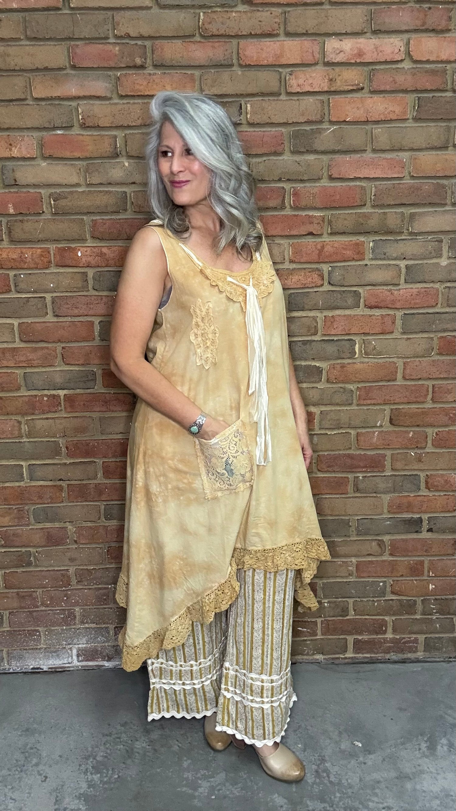 Woman wearing the Wandering Spirit Dress a tea stained tank dress with striped pants standing in front of a brick wall.