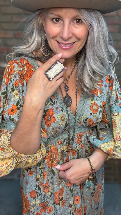 A smiling woman with long gray hair and a wide-brim hat wearing a carved Owl Ring,  a bohemian-style floral dress with orange, blue, and beige tones.