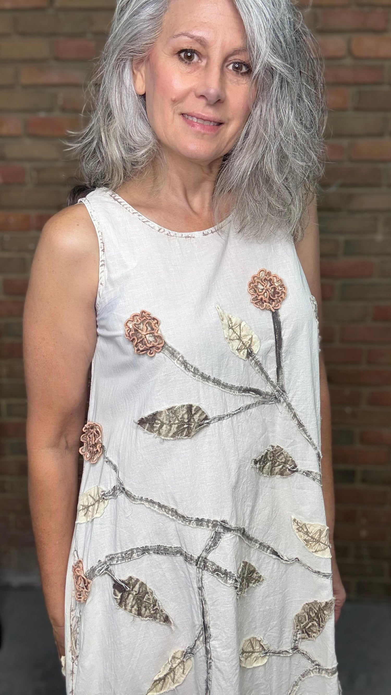 Woman wearing a white sleeveless dress with floral patterns against a brick wall.
