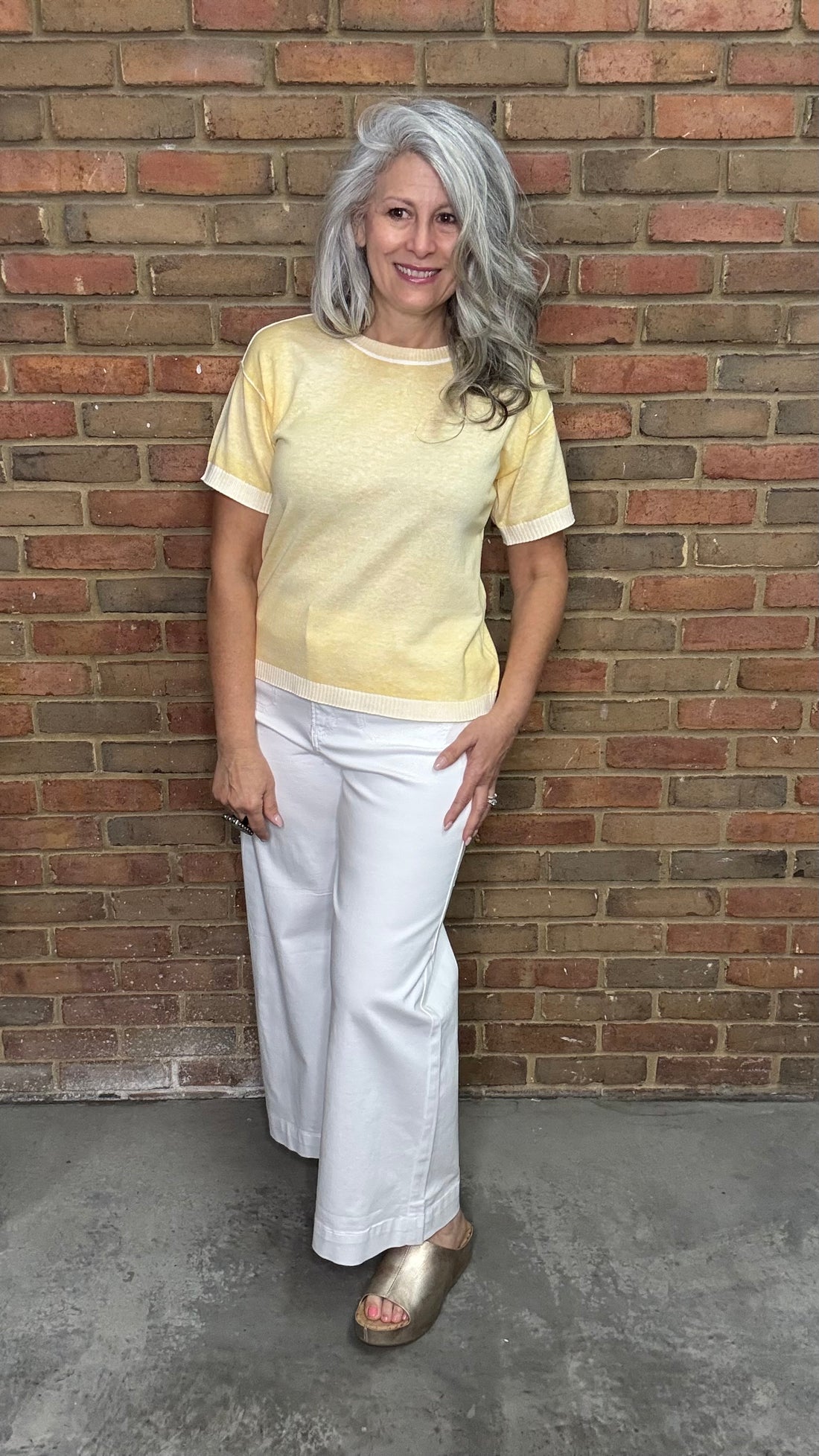Woman wearing the Sunshine Short Sleeve Sweater, a yellow sweater and the White Twill Cropped Flare Leg pants standing against a brick wall.