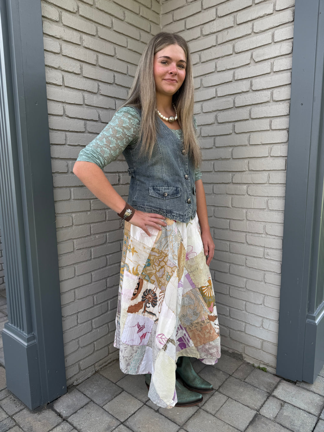 Woman standing against a brick wall wearing a denim vest, patterned skirt, and teal boots.