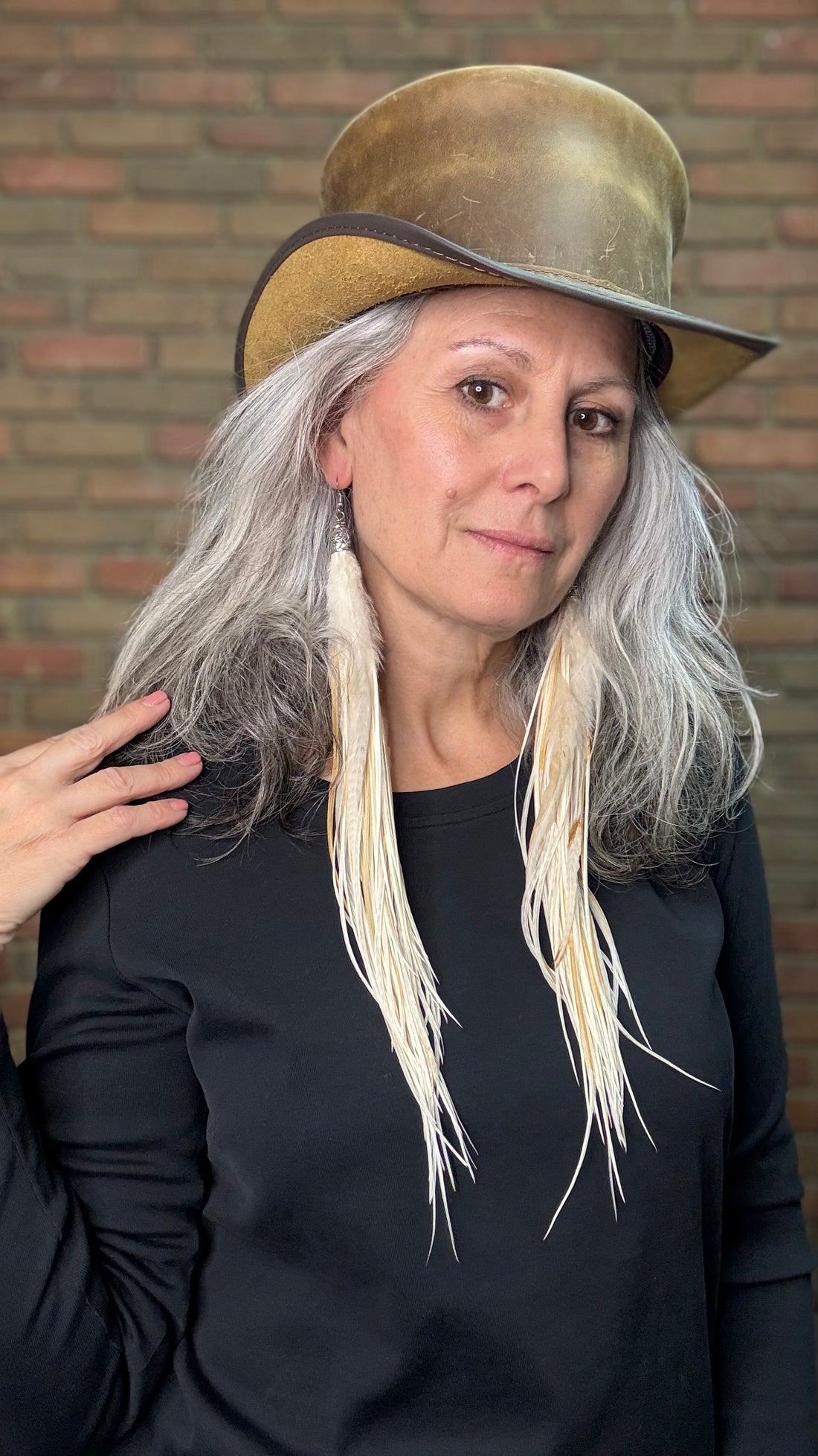 Woman with long feather earrings wearing a brown leather top hat against a brick wall