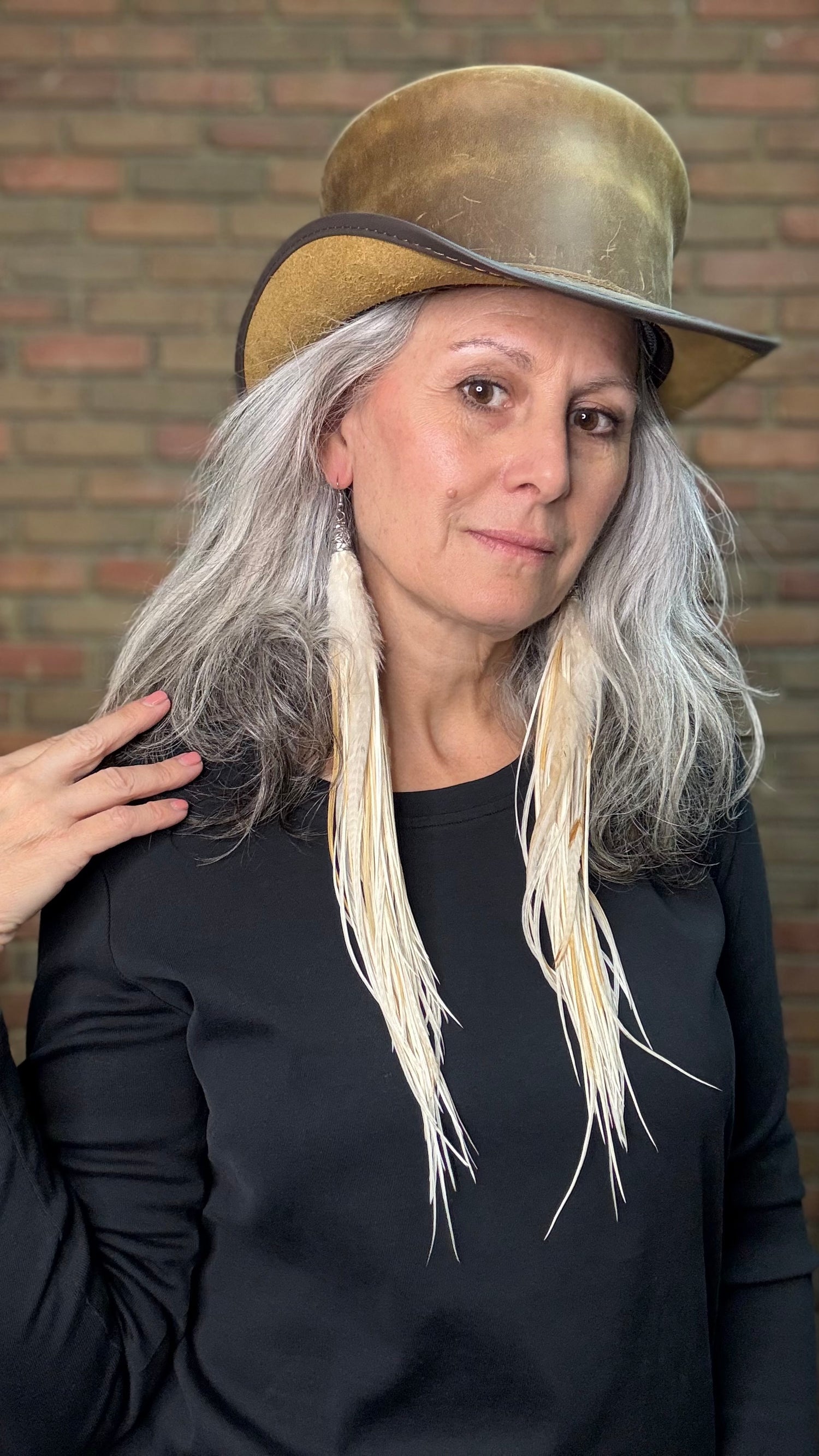 Woman with long feather earrings wearing a brown leather top hat against a brick wall