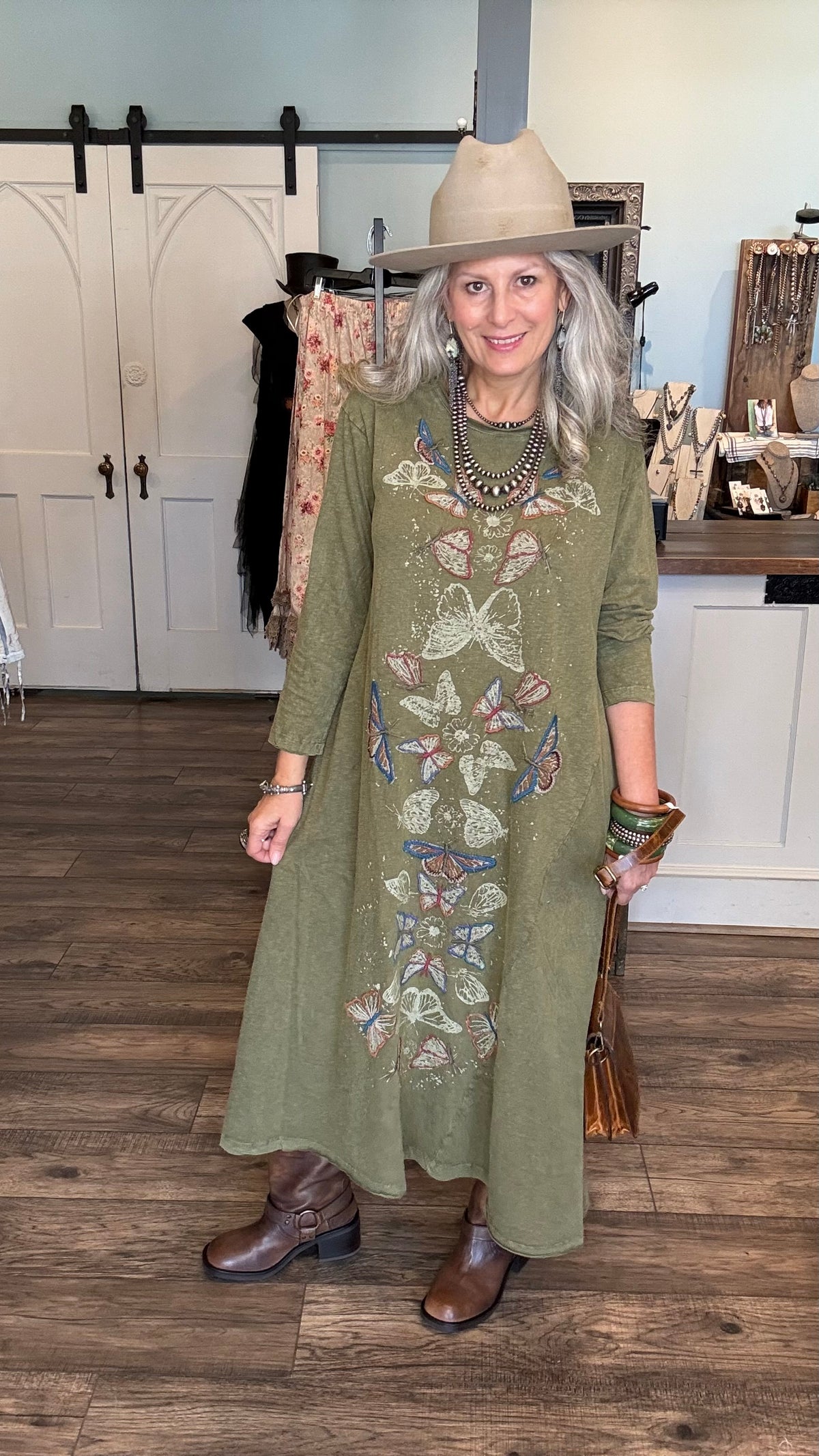 A woman wearing a sage green long-sleeve midi dress with colorful butterfly embroidery, styled with a beige wide-brim hat, brown leather boots, and layered necklaces, standing in a boutique setting.