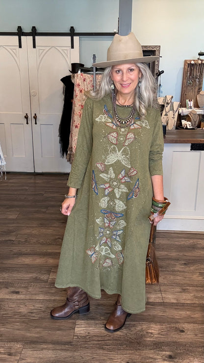 A woman wearing a sage green long-sleeve midi dress with colorful butterfly embroidery, styled with a beige wide-brim hat, brown leather boots, and layered necklaces, standing in a boutique setting.