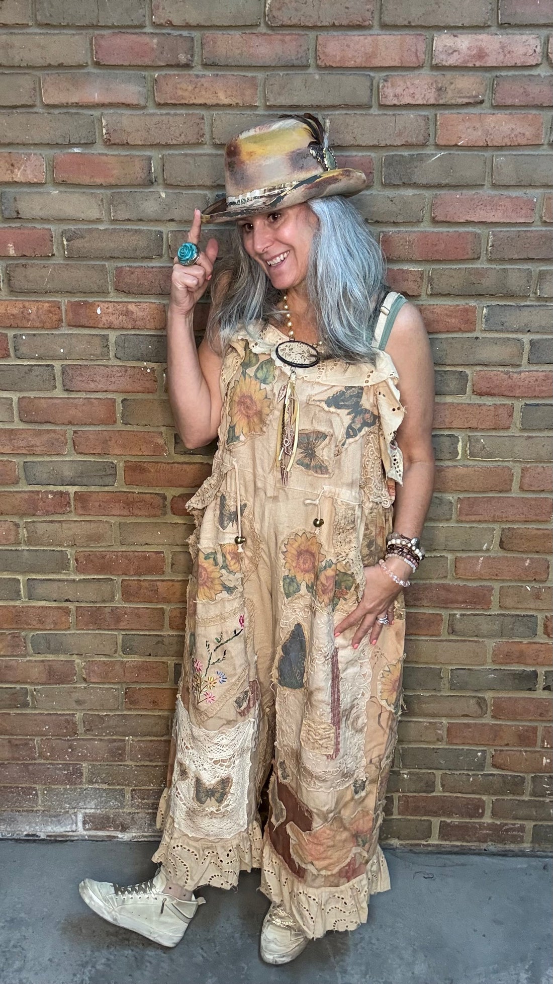 A woman with gray hair wearing a bohemian-style printed jumpsuit, a hat, and white sneakers, posing against a brick wall.