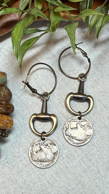 A pair of hoop earrings with antique bronze bottle opener charms and dangling silver coin pendants, displayed on a textured light gray surface with green leaves and brown stones as props.