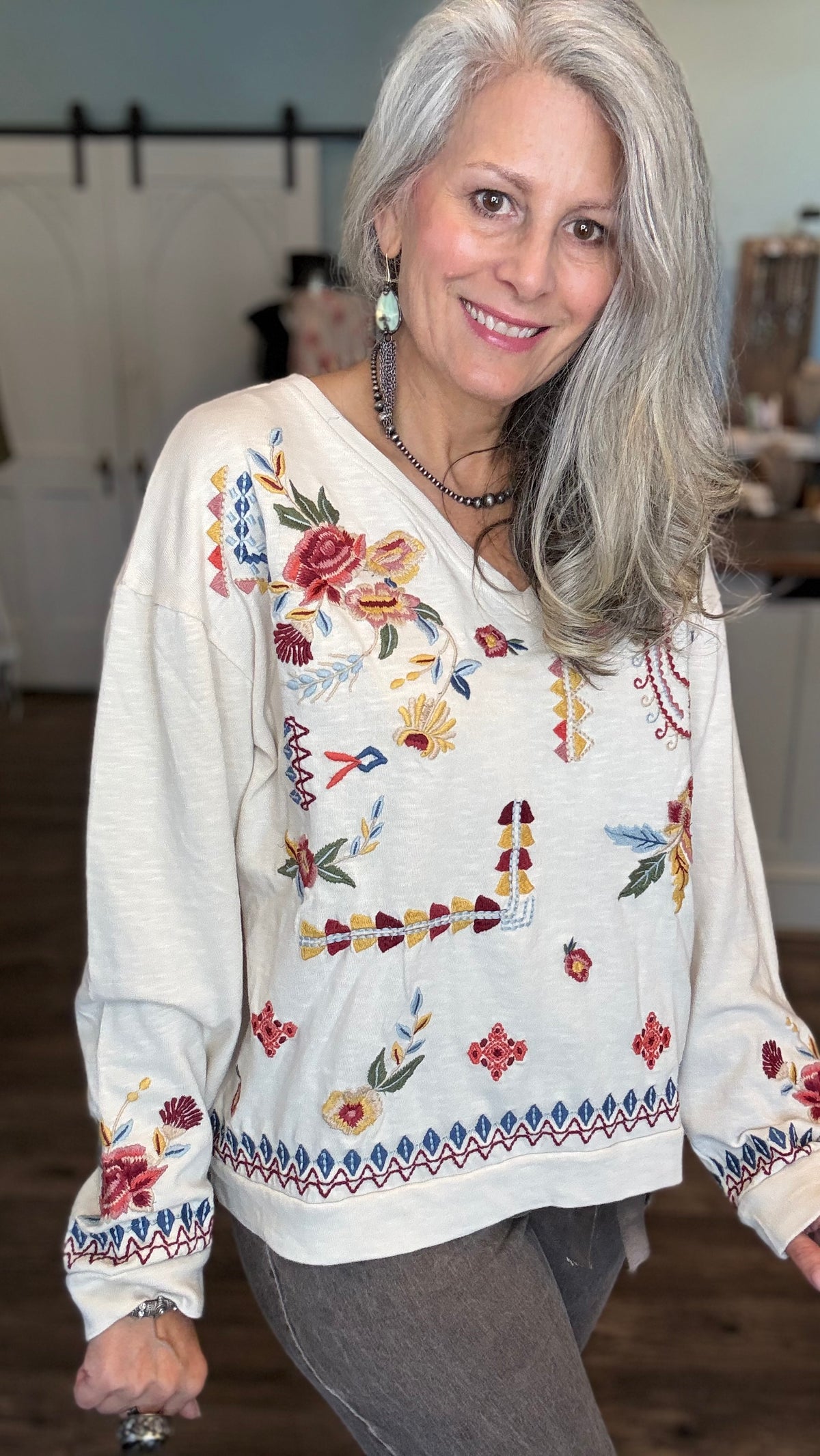 A smiling woman with long gray hair wearing a cream-colored long-sleeve top with colorful floral and geometric embroidery, paired with gray pants, in a softly lit indoor setting.
