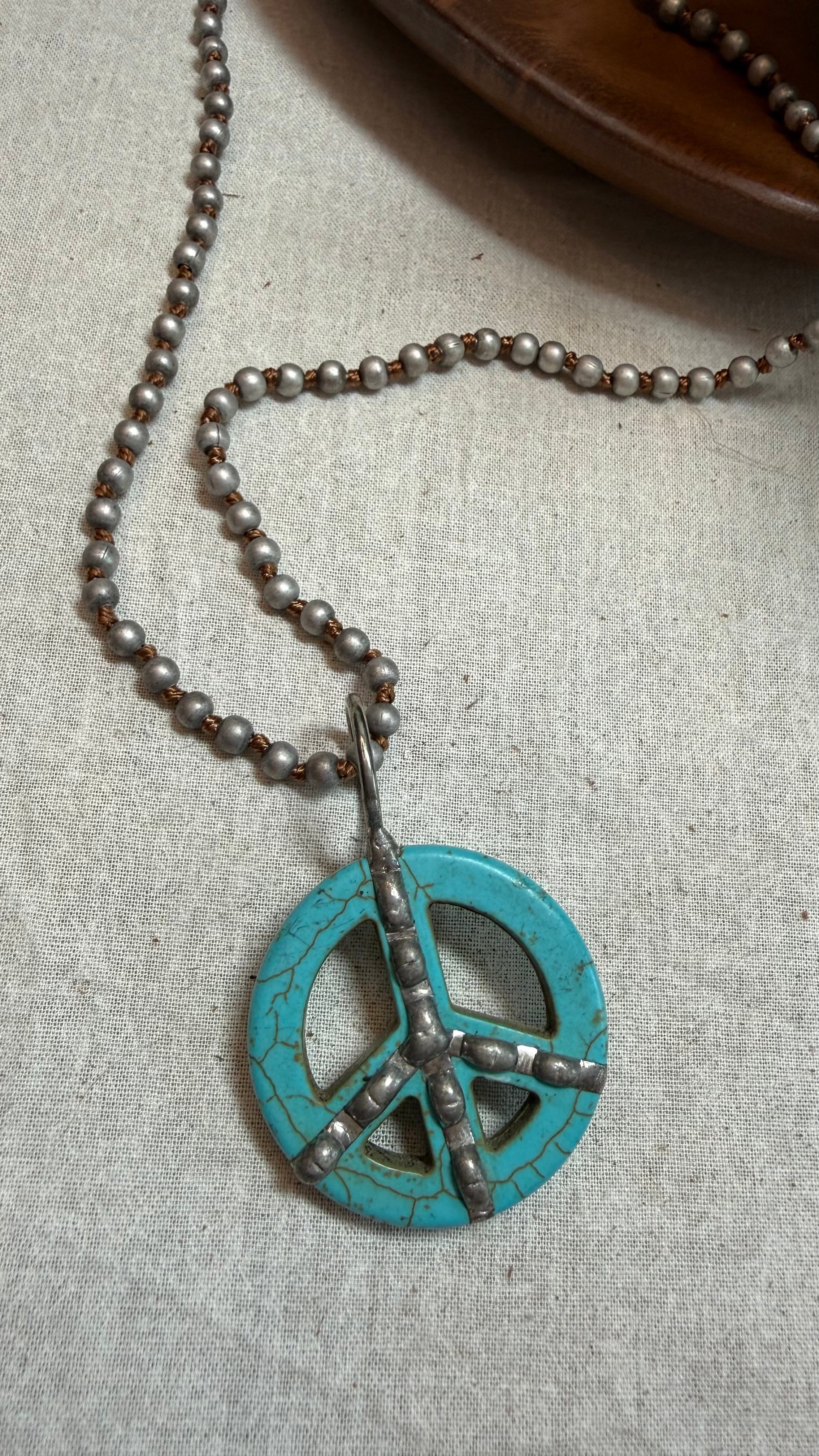 A turquoise and silver peace sign pendant necklace on a beaded chain, displayed on a textured beige linen fabric.