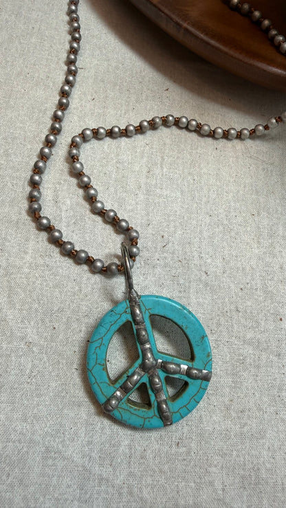 A turquoise and silver peace sign pendant necklace on a beaded chain, displayed on a textured beige linen fabric.