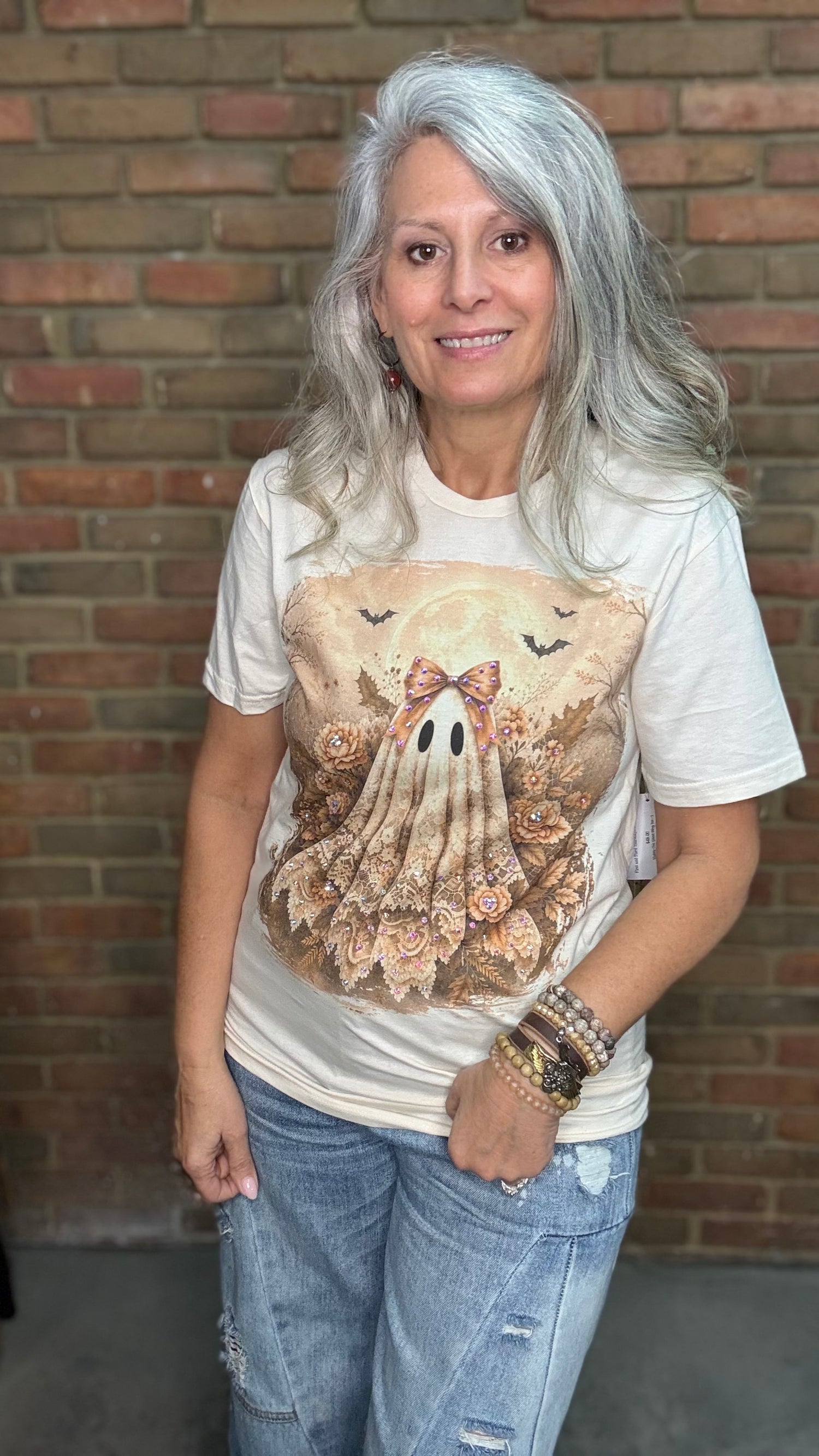 A woman with long gray hair wearing a cream-colored graphic t-shirt with a ghost design and distressed blue jeans, standing in front of a brick wall.