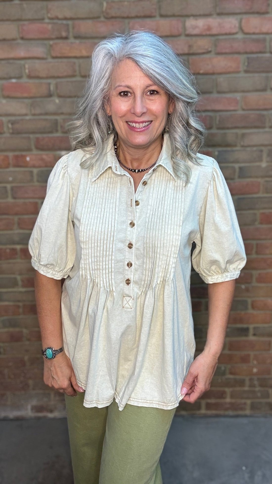 Woman wearing the Khaki Linen Henley Top, a off white pin tucked top and green pants standing in front of a brick wall.