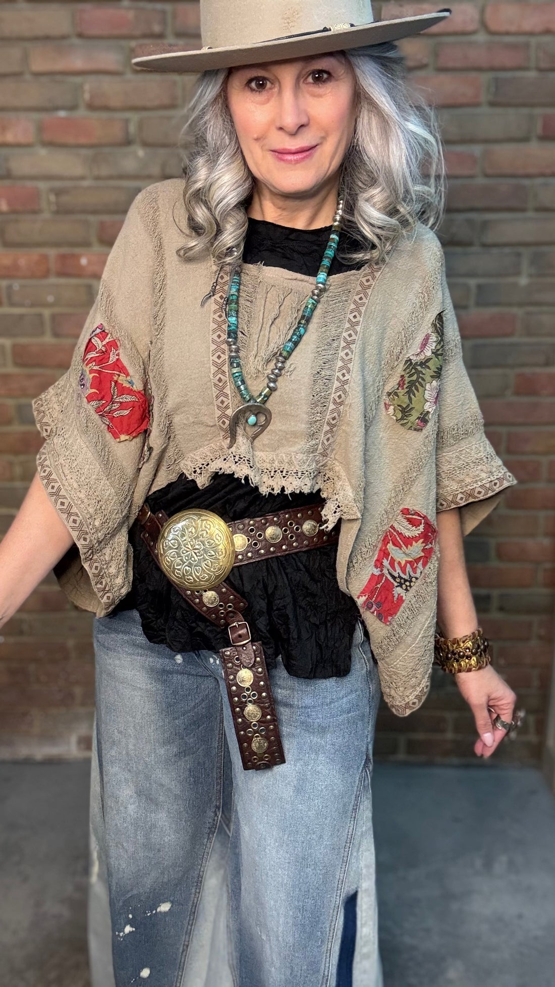 Woman wearing Garden Drift Top with a hat, jeans, and belt against a brick wall.