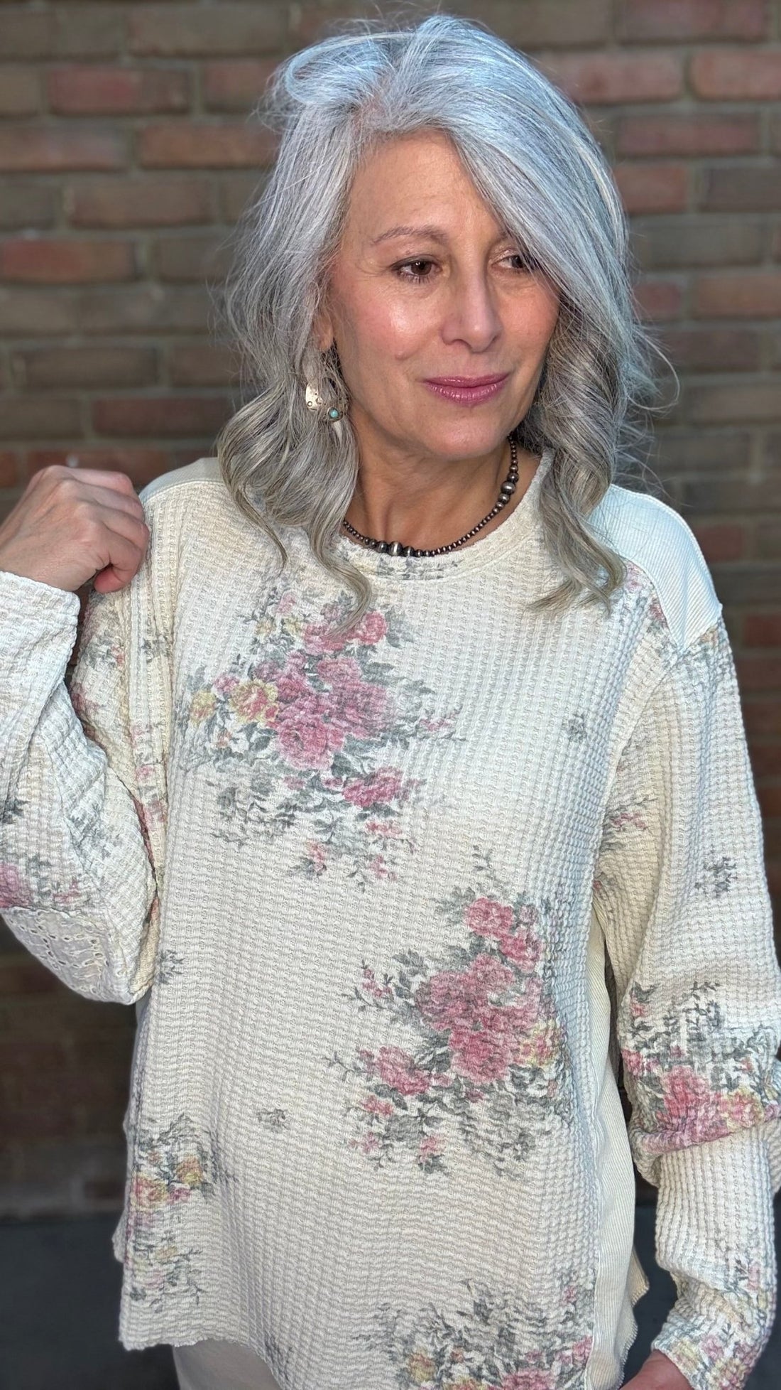 Woman wearing the Waffle Floral Pull On Top, a floral print waffle knit top and beige pants standing against a brick wall.