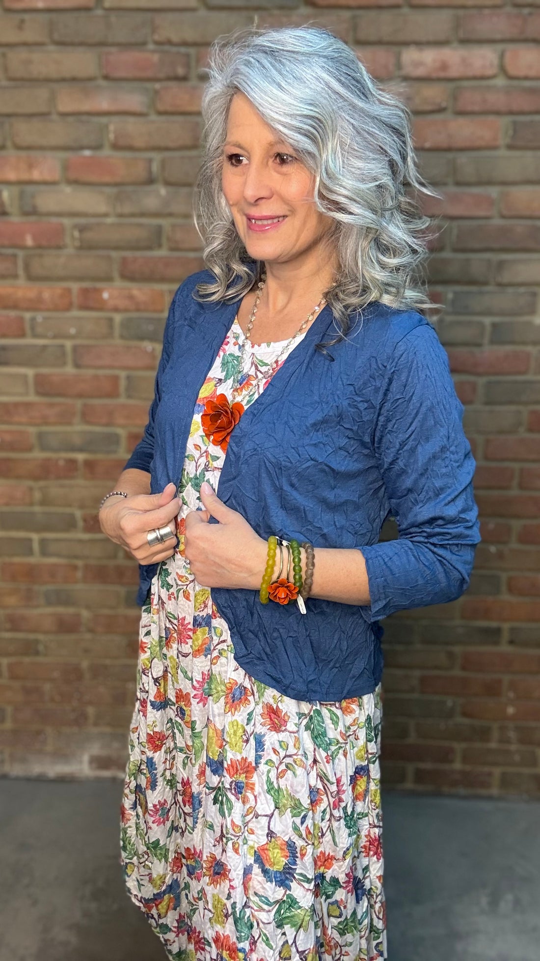Woman with gray hair wearing Indigo Crinkle Cardigan over a floral dress against a brick wall.