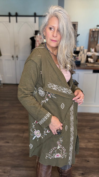 A woman with long silver-gray hair models an olive green embroidered cardigan in a boutique setting. The cardigan features white geometric patterns and floral embroidery with pink and purple accents.